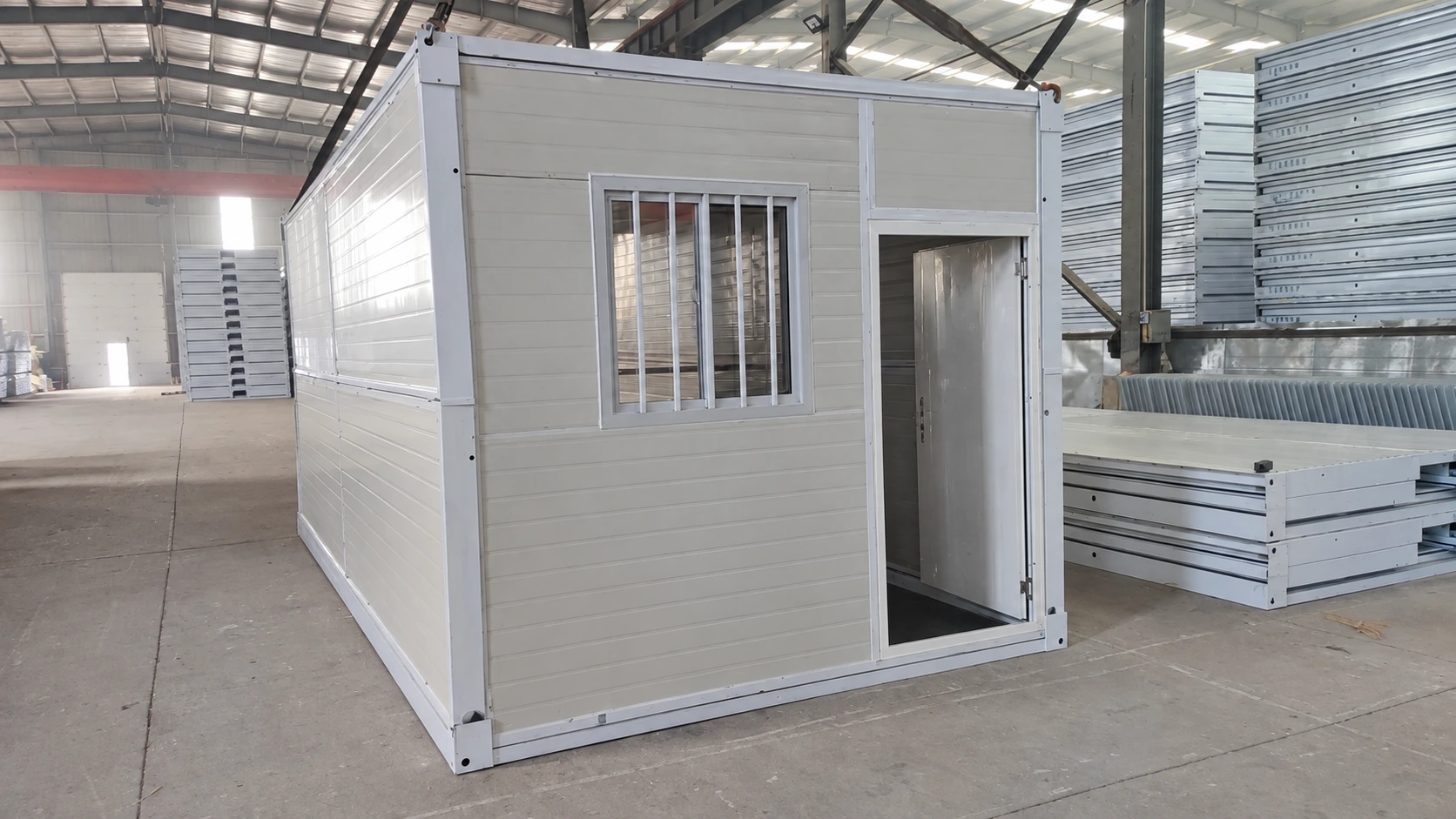 En la imagen de referencia del taller de producción se muestra una habitación modular gris ya ensamblada, con una ventana enrejada y una puerta abierta.