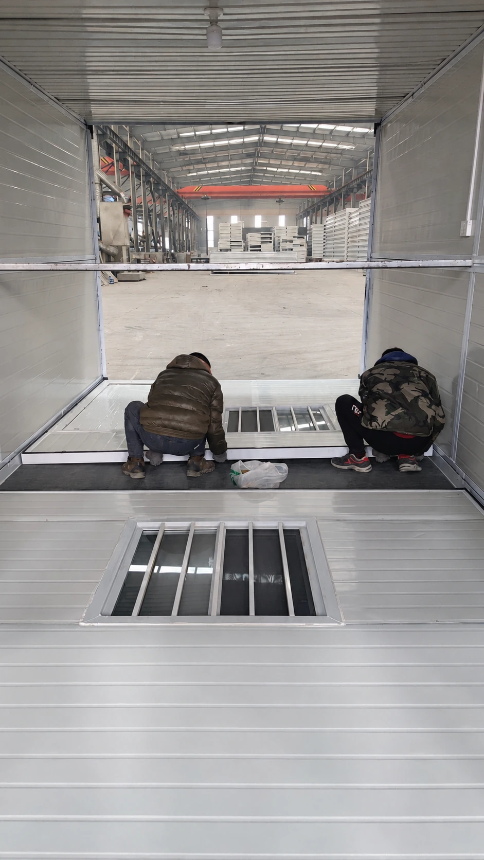 En la imagen se muestra un taller de producción con dos trabajadores en el montaje de un panel de pared plegable dentro de un módulo abierto.
