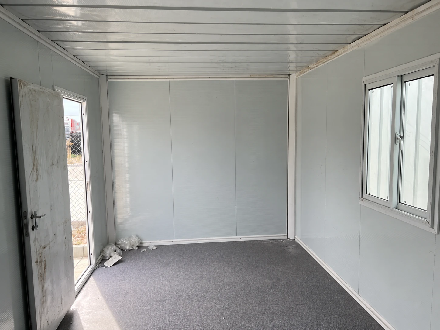 Leerer paneelverkleideter Raum mit offener Tür links, Schiebefenster rechts und grauem Boden. Referenz für Speiseraumnutzung.