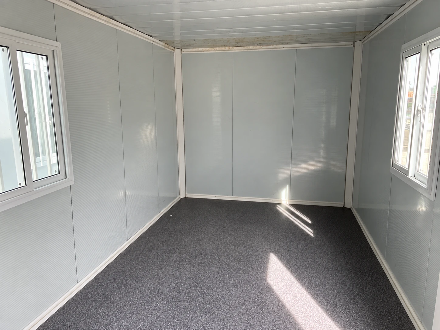 Leerer paneelverkleideter Raum mit gegenüberliegenden Fenstern, Metallpaneeldecke und grauem Boden. Referenz für Speiseraumnutzung.