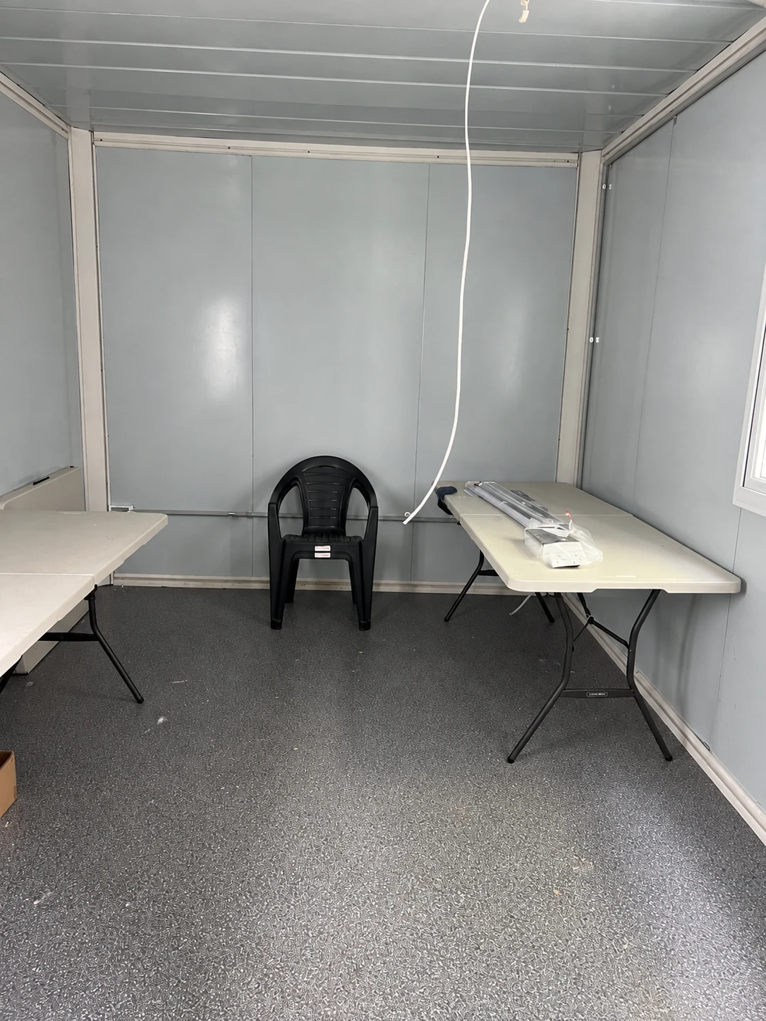 Klapptische an beiden Seiten eines paneelverkleideten Raums, schwarzer Stuhl, verpackte Leuchte und loses Deckenkabel. Referenz für Speiseraumnutzung.