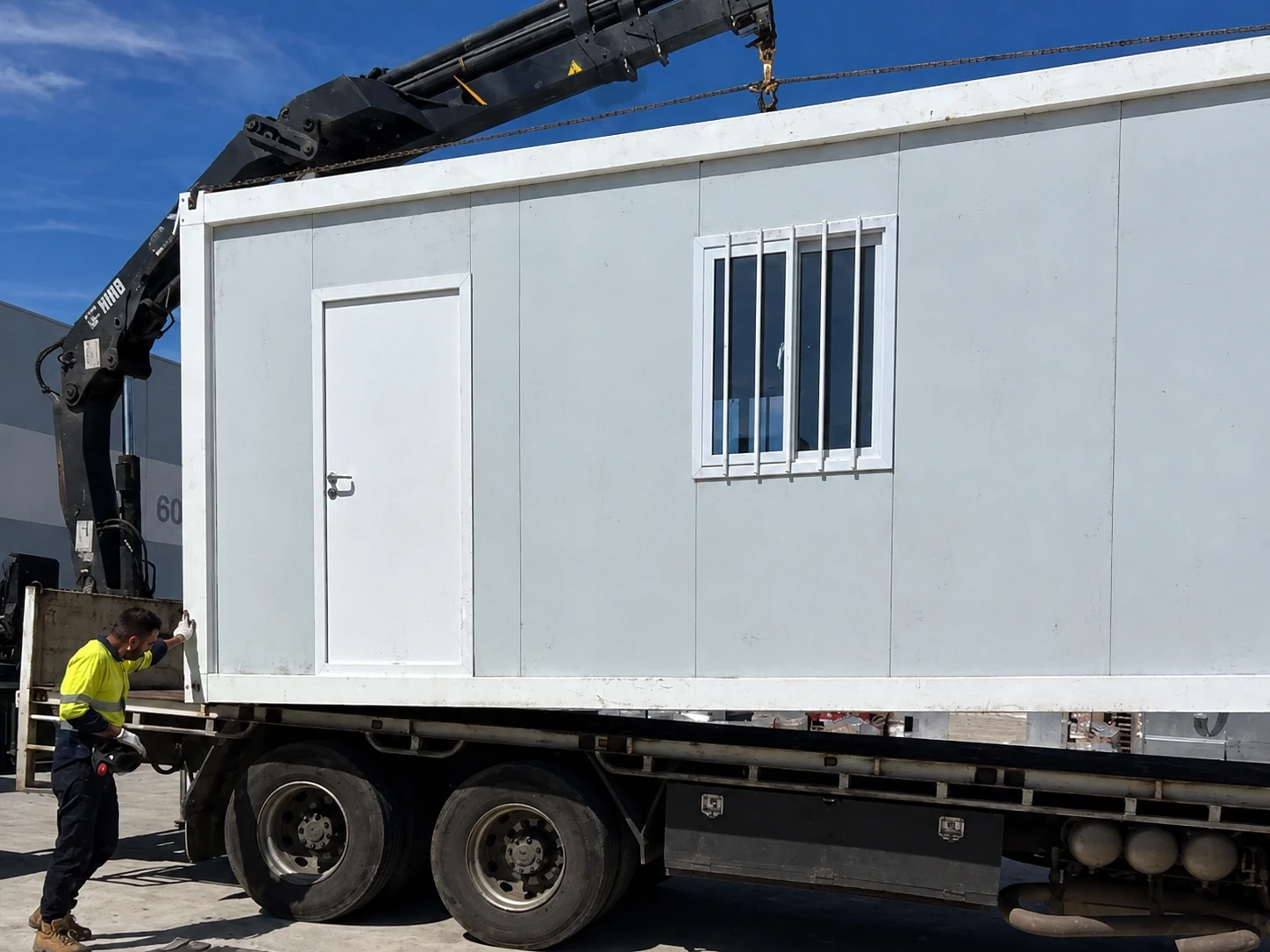 Weißes Modul mit Tür und vergittertem Fenster auf einem Pritschen-Lkw unter einem Kranausleger, mit Arbeiter an der Ecke. Referenz für Speiseraumtransport.