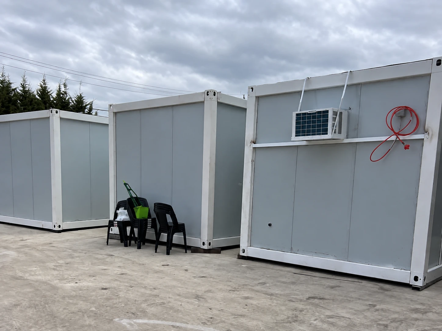 Weiße Paneelmodule auf einer Betonfläche mit Fensterklimagerät und gestapelten schwarzen Stühlen. Referenz für Speiseraumnutzung.