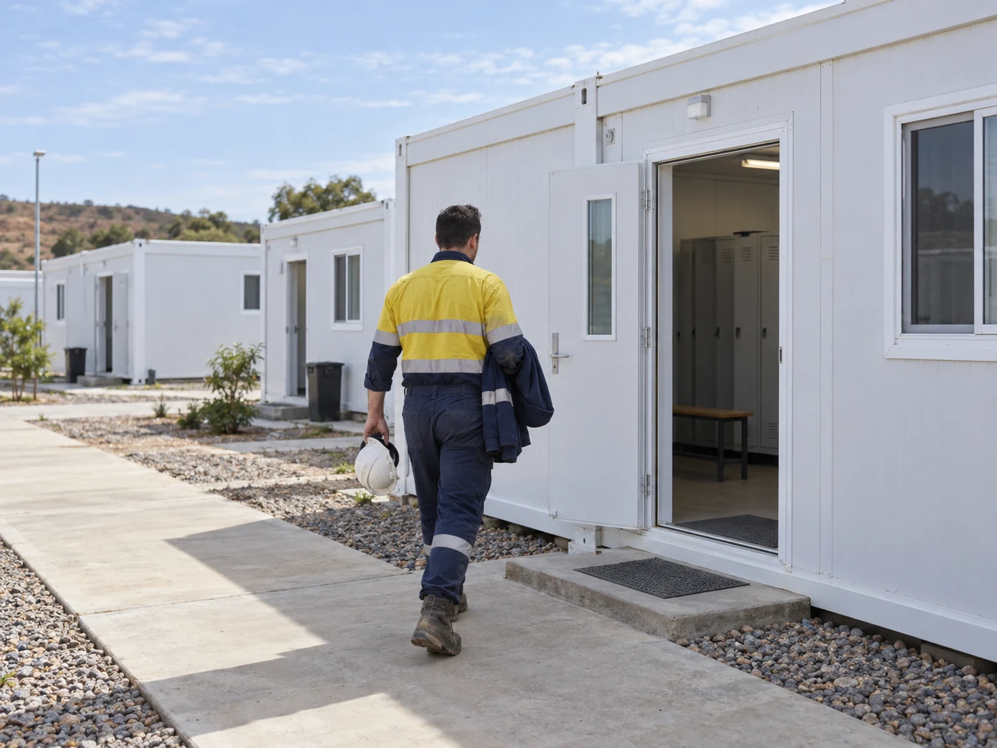 A worker carrying a helmet and jacket toward a changing module along a modular camp path