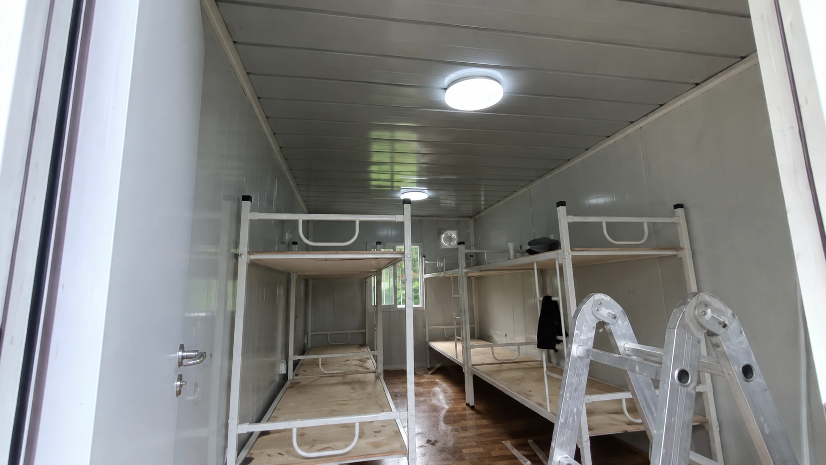 Etagenbettgestelle auf beiden Seiten eines schmalen paneelverkleideten Raums mit Leiter, Deckenleuchten und hinterem Fenster. Allgemeine Innenraumreferenz.