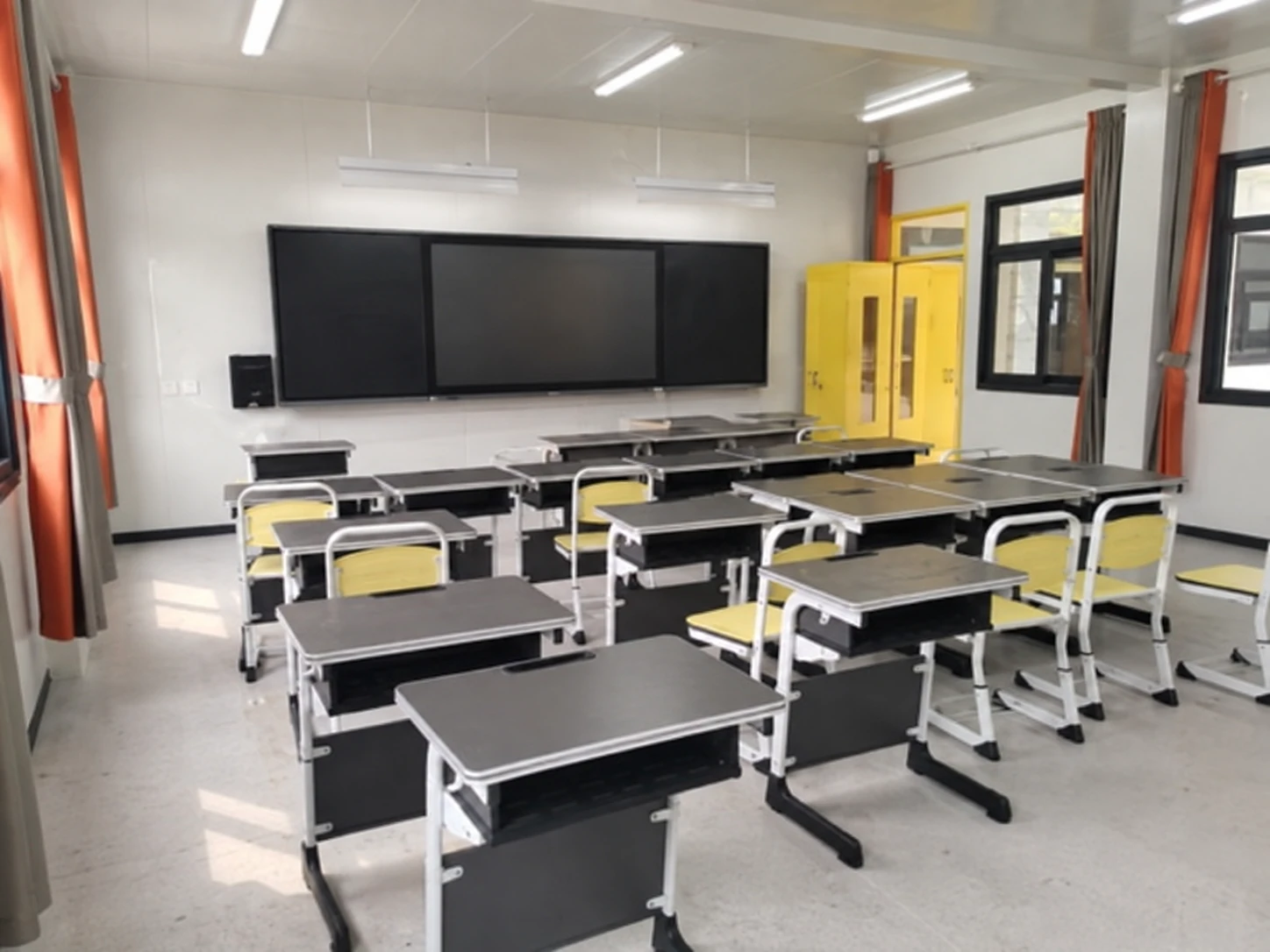Rows of classroom desks and chairs facing a large front-wall display inside a modular room. Shared classroom-layout reference.