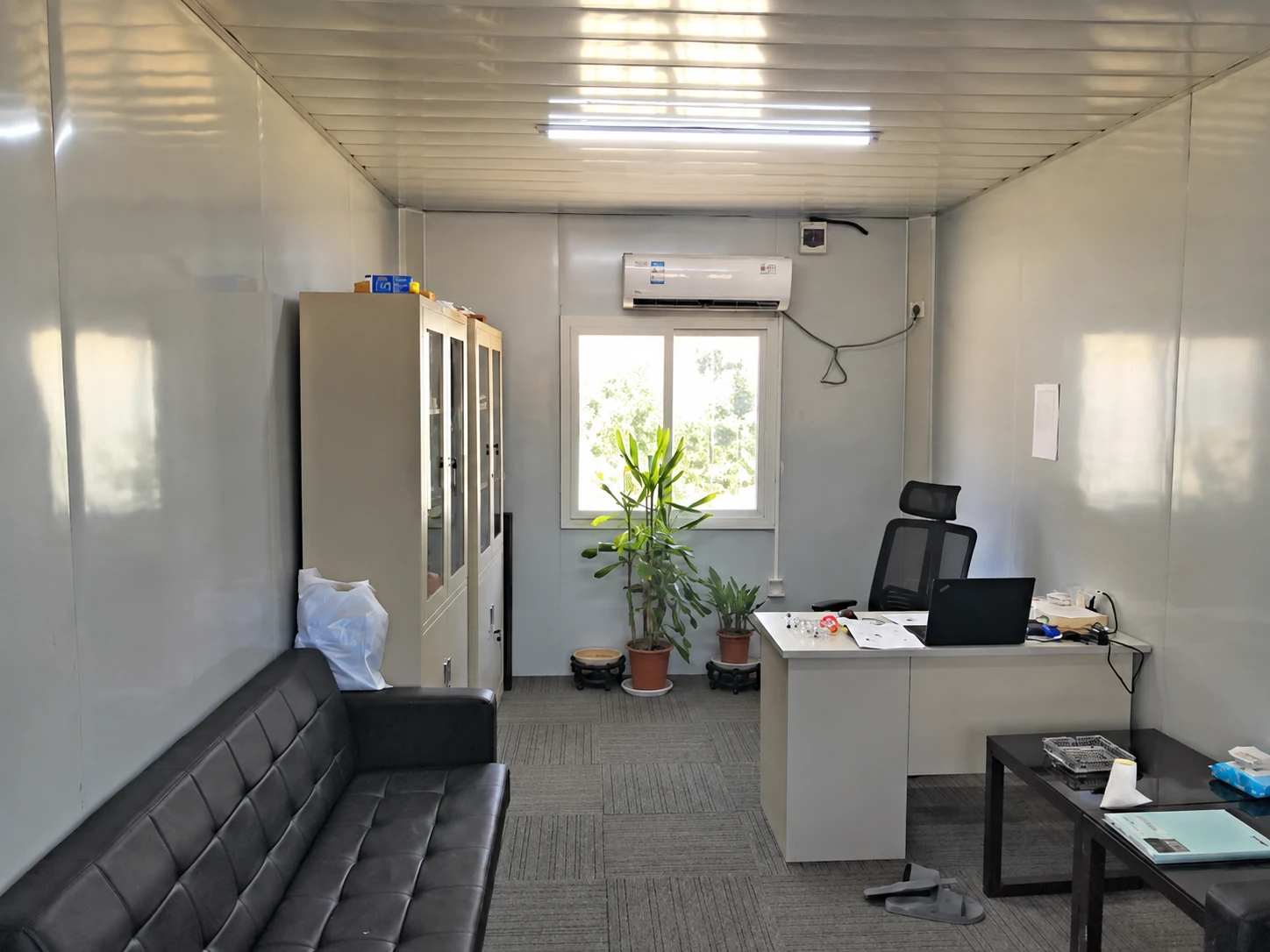 Kompaktes paneelverkleidetes Büro mit Schreibtisch, Laptop, schwarzem Sofa, hohen Schränken und Klimagerät über einem Fenster. Allgemeine Innenraumreferenz.