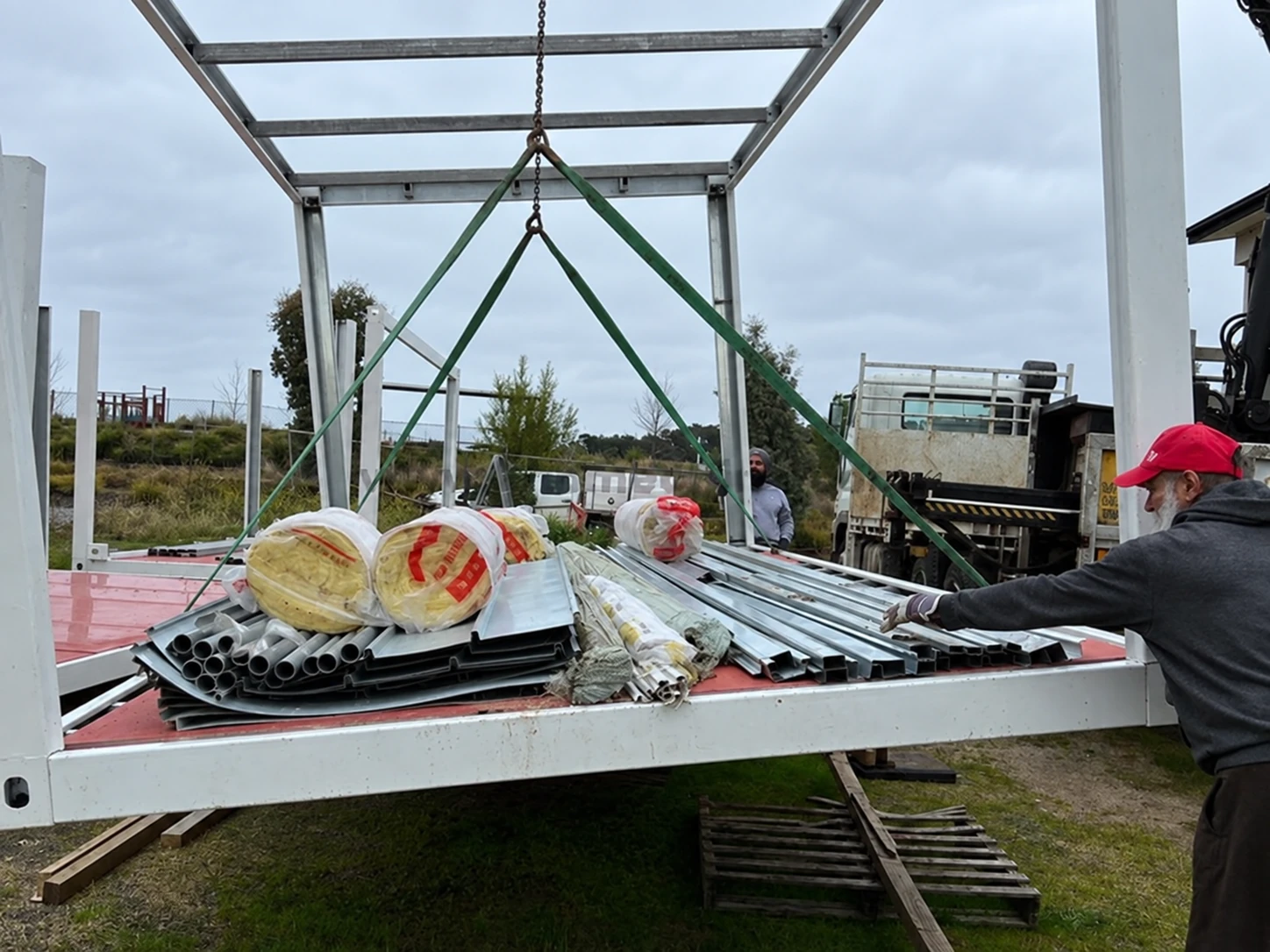 Offener Modulrahmen mit Rohren, Metallprofilen und verpackter Dämmung, grünen Hebegurten sowie nahegelegenem Lkw und Arbeitern. Montagereferenz.