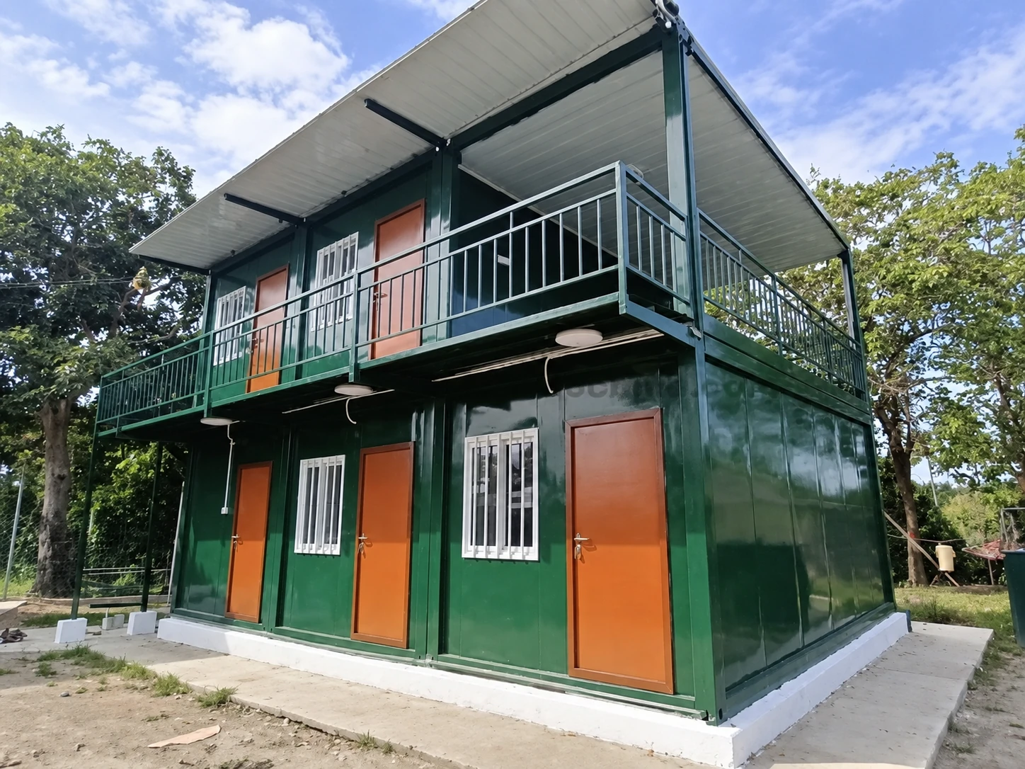 Grünes zweigeschossiges Modulgebäude mit orangefarbenen Türen, vergitterten Fenstern, oberem Balkon und breitem Vordach. Allgemeine Außenreferenz.