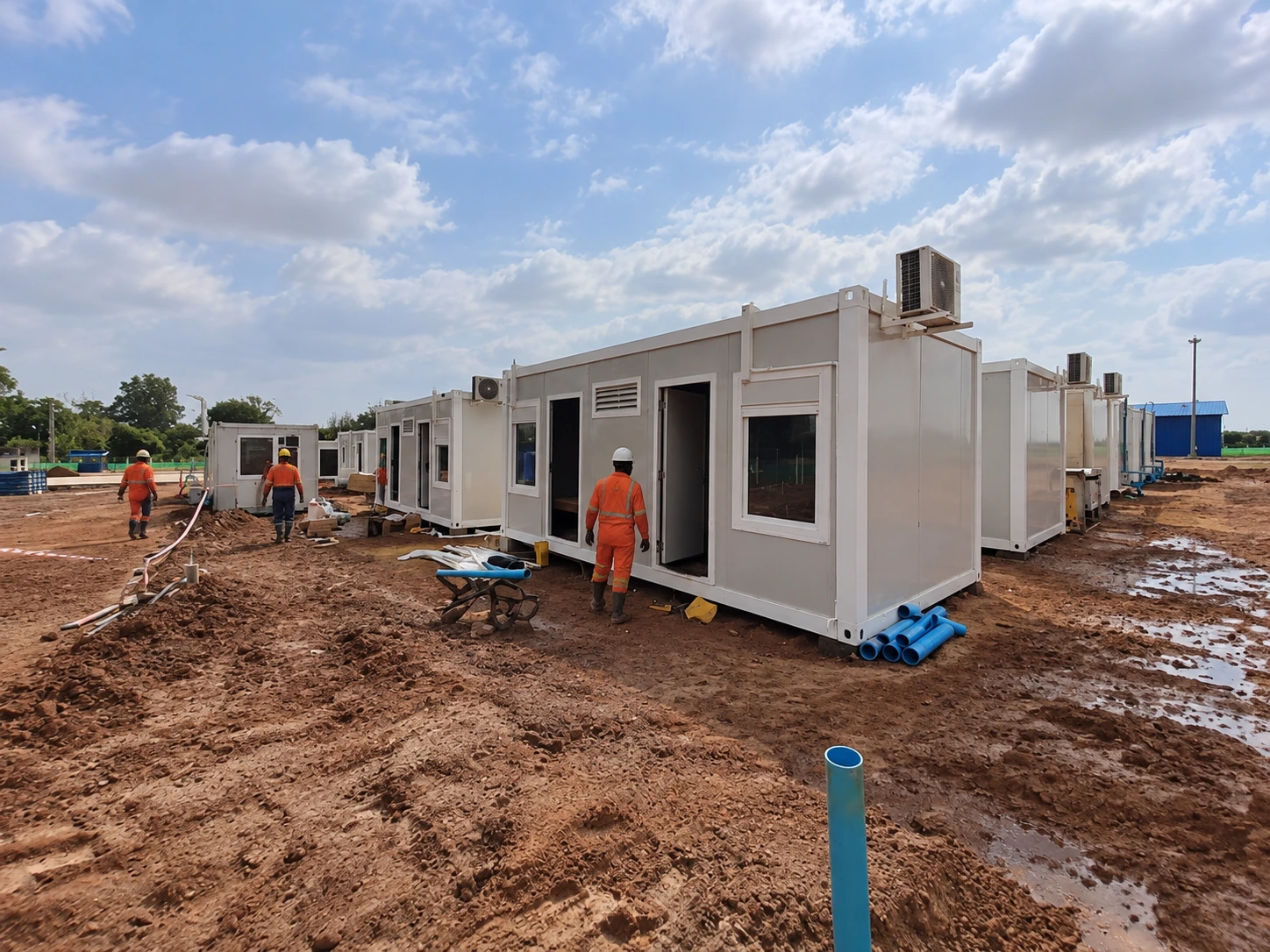 Foto de referencia de la instalación: habitaciones modulares de una sola planta, trabajadores, carretillas y tomas de servicios expuestas junto al suelo desnudo.