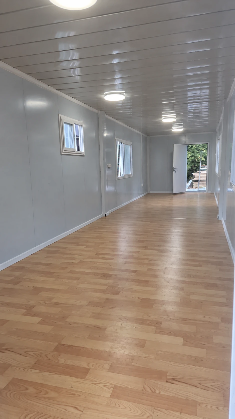 Long unfurnished room with wood-look flooring, white panel walls, small side windows and an open door at the far end. Interior layout reference.