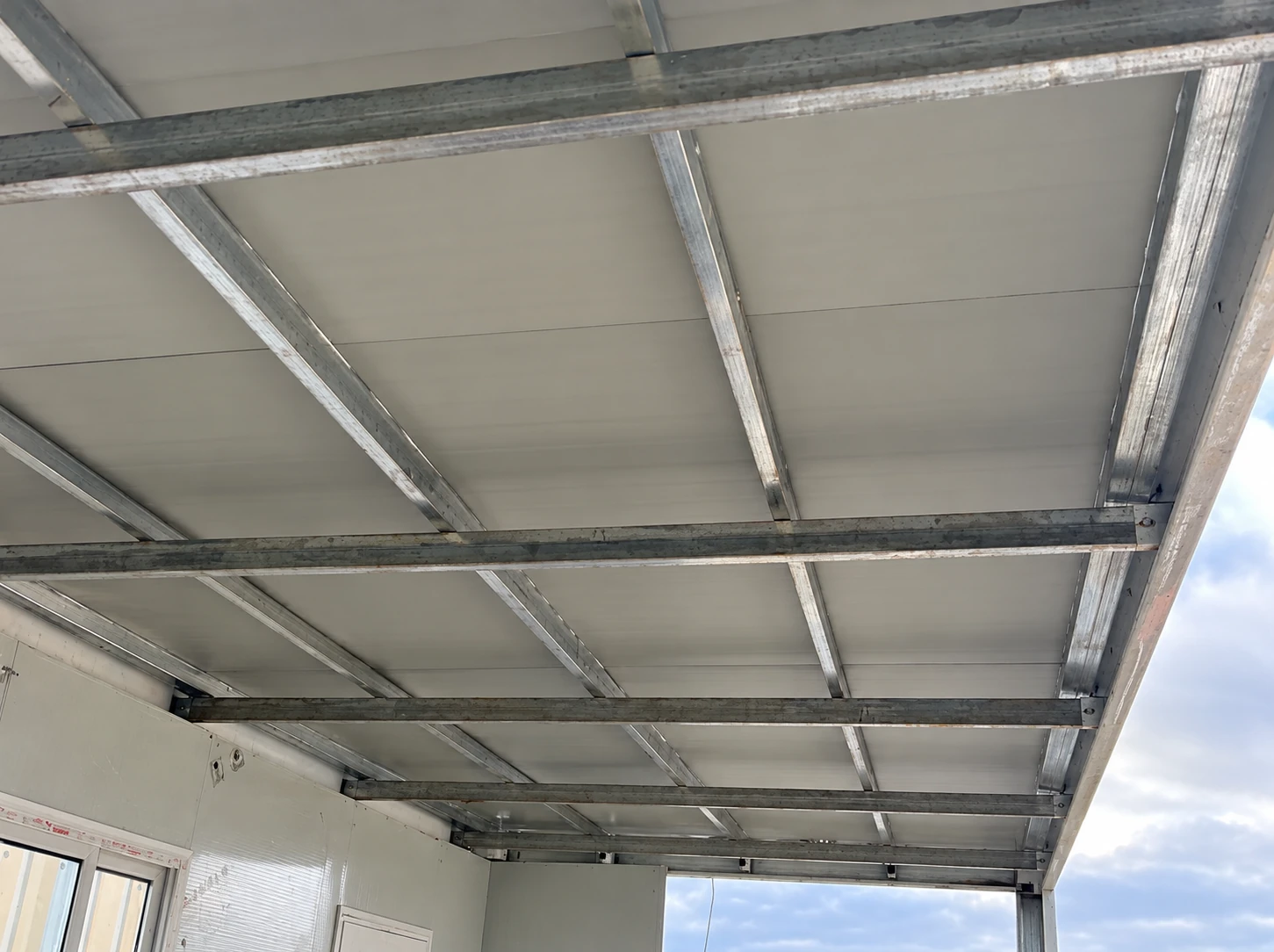 Unterseite von Dachpaneelen auf Metallquerträgern über einem teilweise verkleideten Modul mit Fenster. Referenz für Montagedetails.
