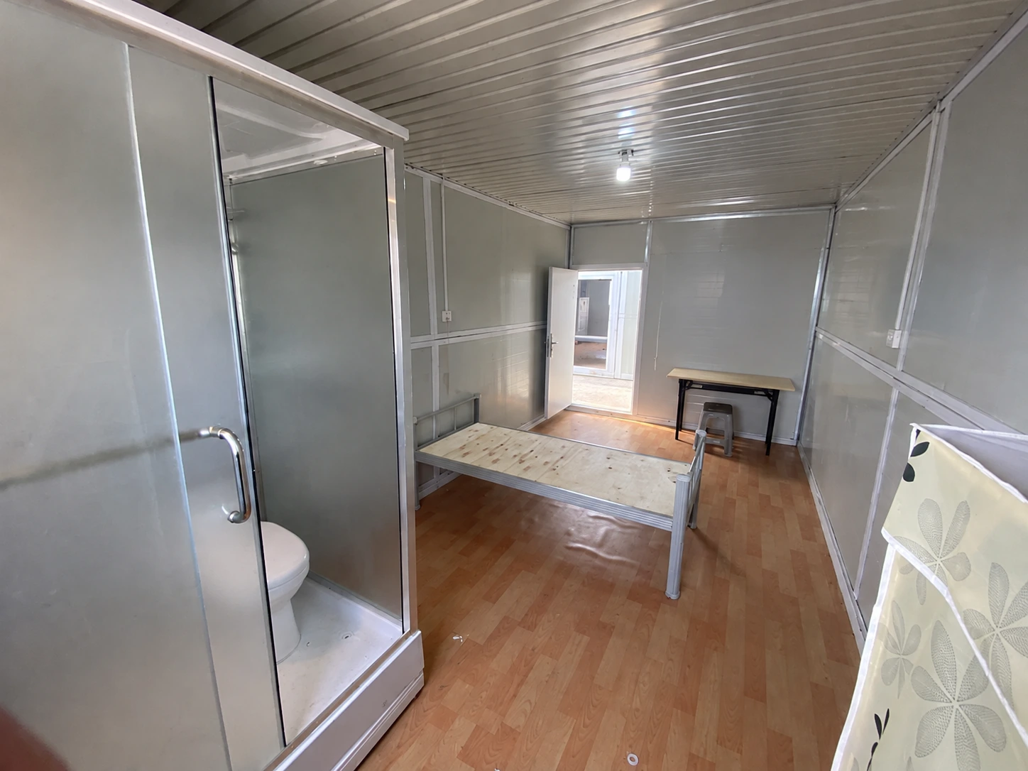 Foto de referencia del interior de una habitación modular con estructura de cama, escritorio pequeño y área de aseo separada.