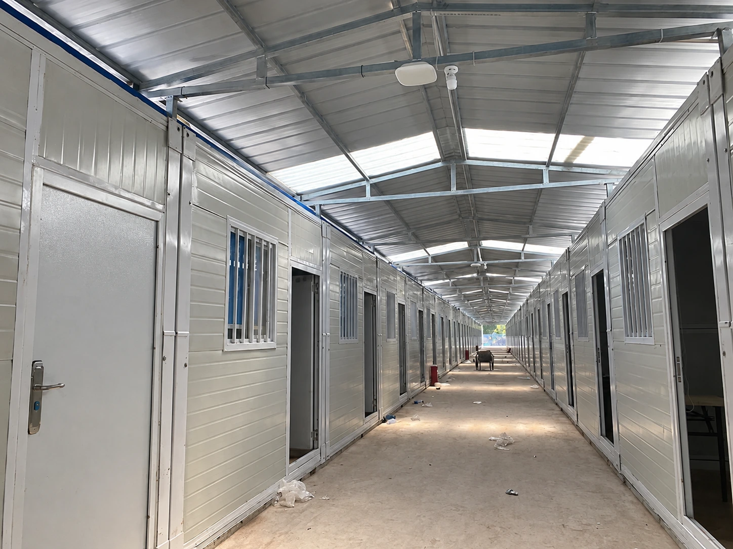 Foto de referencia de un pasillo cubierto entre habitaciones modulares, con puertas abiertas, ventanas enrejadas y luces en el techo.