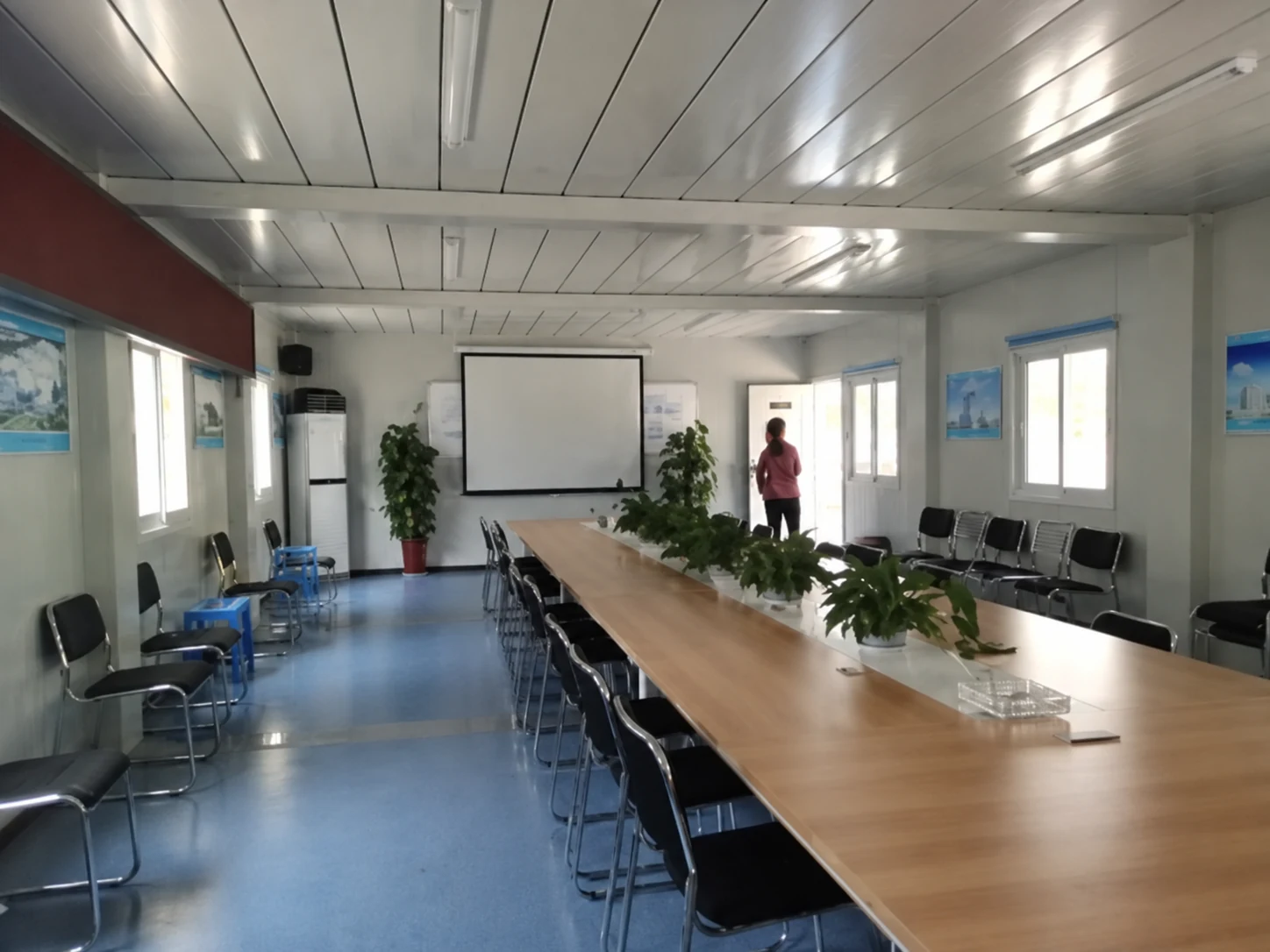 Long meeting table, rows of chairs and a wall screen inside a modular room; wider room-use reference.