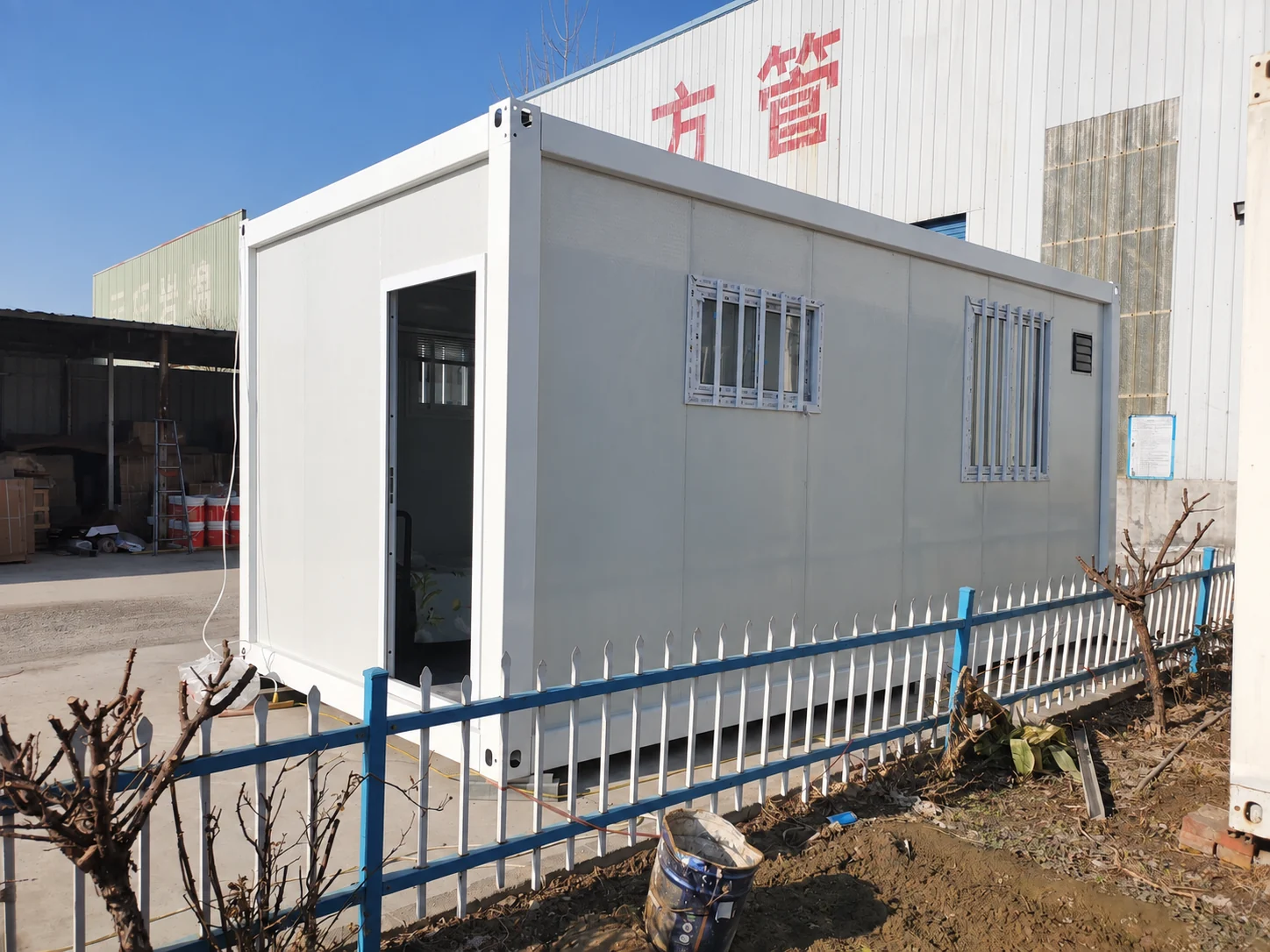 Module de pièce blanche avec entrée ouverte et fenêtres latérales grillagées, jouxtant un bâtiment industriel. Référence de fabrication.