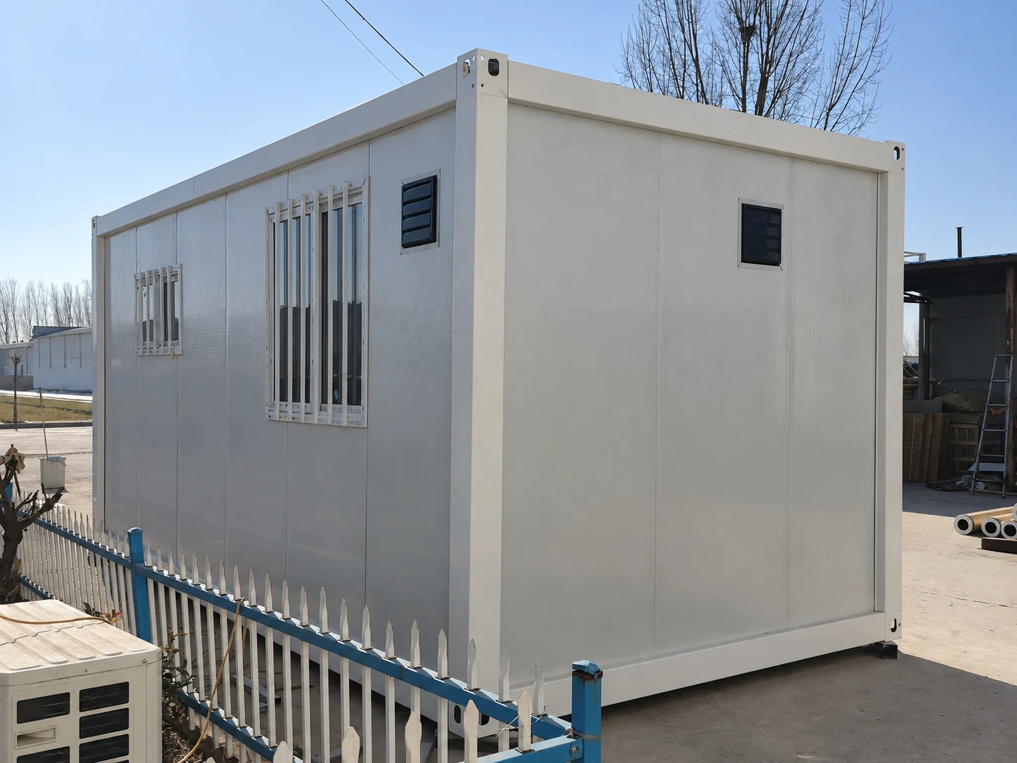Vues arrière et latérale d'un module de pièce blanche avec fenêtres à barreaux et petites grilles d'aération murales dans une cour d'usine. Référence de fabrication.