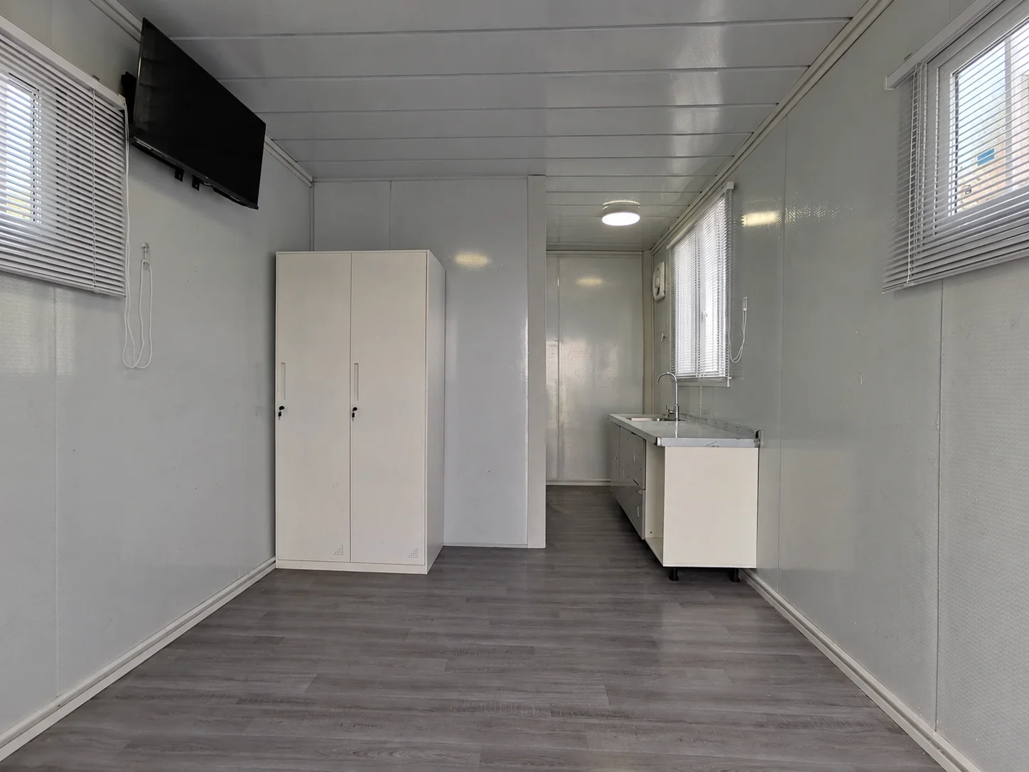 Pièce témoin non meublée avec parquet gris, armoire, meuble-lavabo, stores et télévision murale. Exemple d'aménagement intérieur.