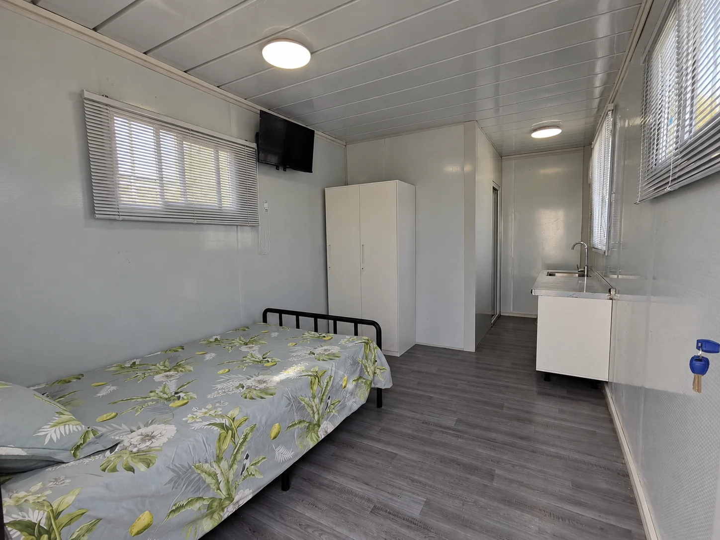 Aménagement intérieur type d'une chambre avec un lit simple, une armoire blanche, un meuble-lavabo, des stores et une télévision murale. Référence d'aménagement intérieur.