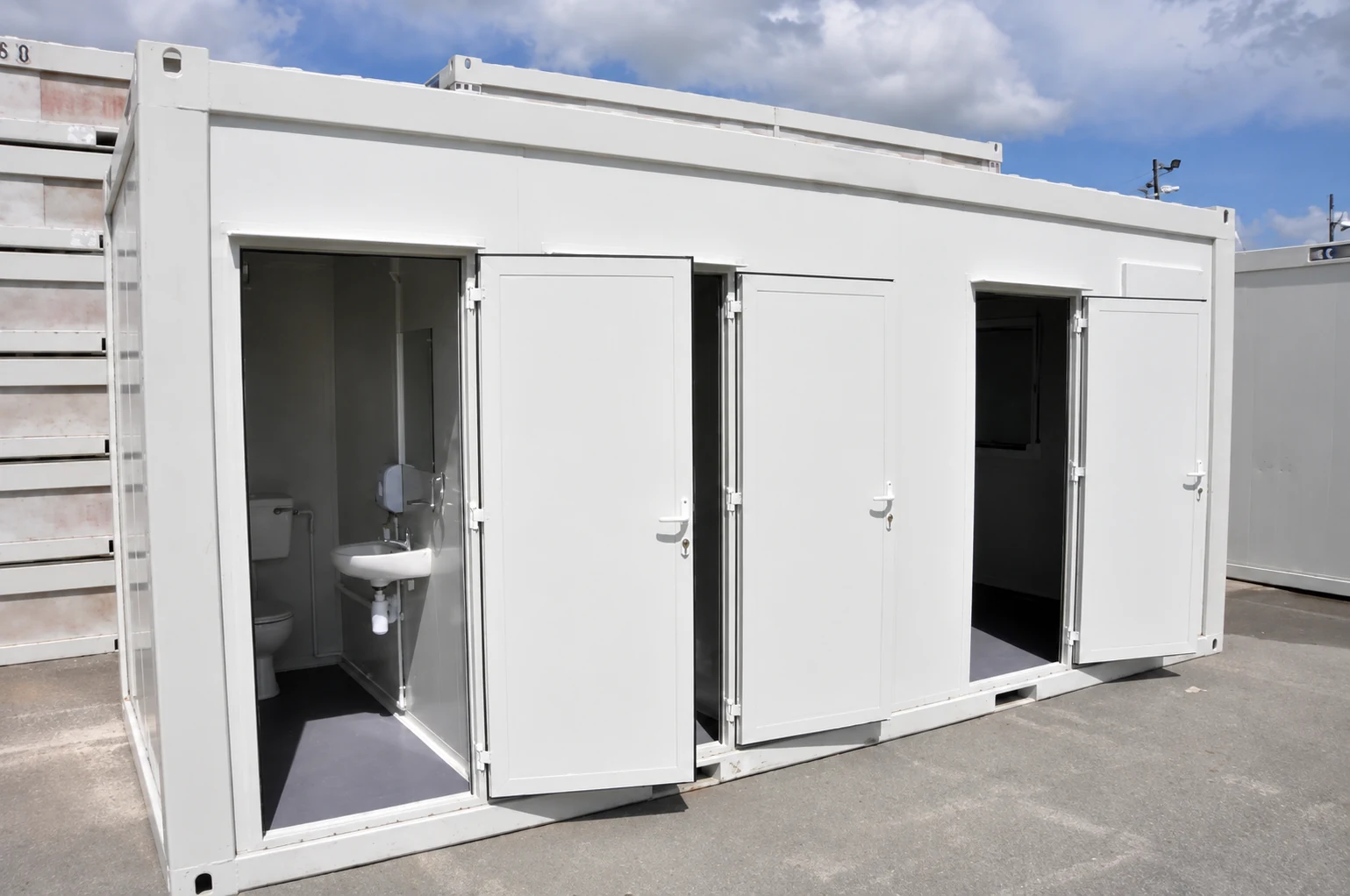 Open doors reveal toilet cubicles and washbasins in a white portable sanitation block; product reference.