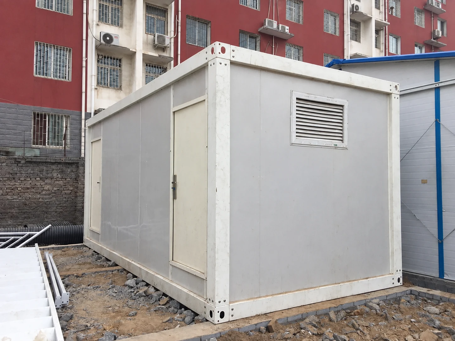 Small white portable toilet cubicle with a door and louvred wall vent in a yard; product reference.