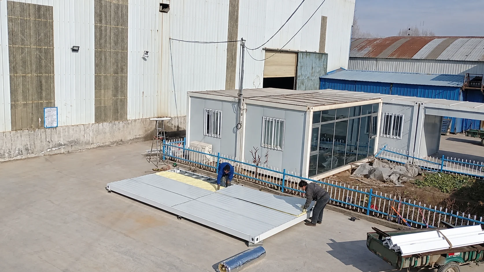Ouvriers assemblant une longue base de plancher près de modules blancs de chambres et d’un bâtiment d’usine. Référence de fabrication.