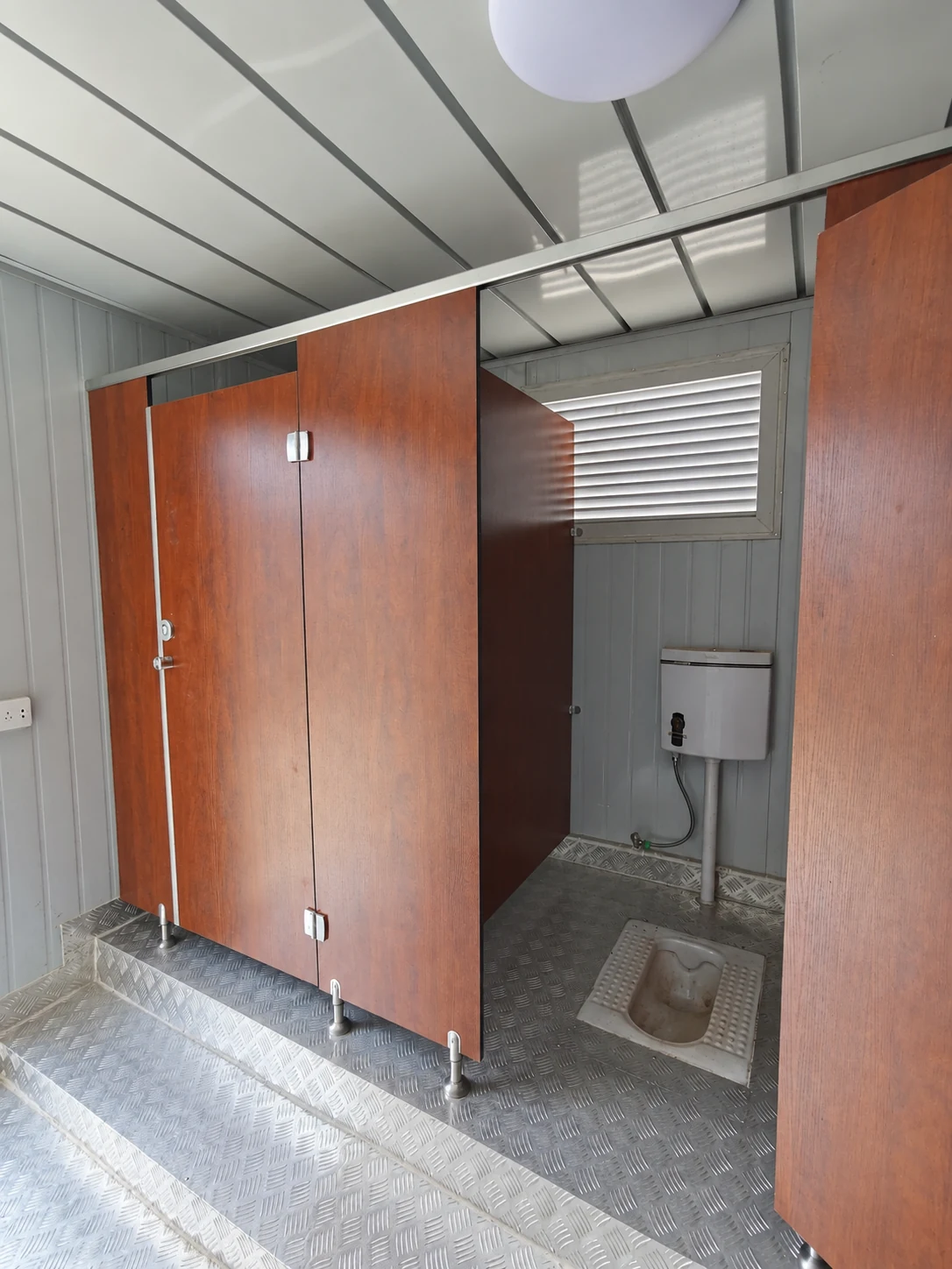 Interior reference showing a squat toilet pan, brown partition door, wall vent and raised floor edge.