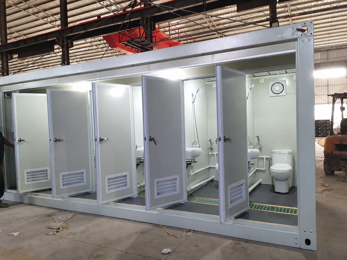 Open wet-room cubicles with toilets, basins, shower fittings and vents inside a production workshop; manufacturing reference.