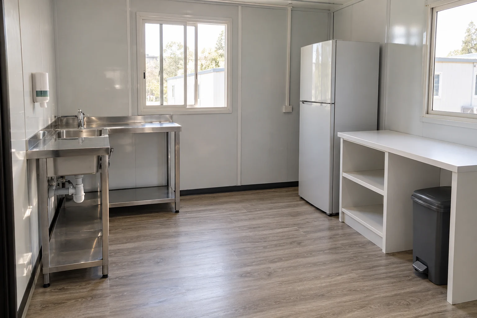 Un fregadero de acero inoxidable, una papelera cerrada independiente con pedal y un refrigerador cerrado ocupan posiciones distintas en una pequeña cocina sencilla con paredes de paneles.